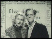 En ung man håller armen kring en kvinna i samma ålder, tidningsnotis i bakgrunden.