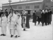 Präst med korgossar, i bakgrunden män som bär på en kista. Det hela utspelar sig på Bromma flygplats och det är den italienske ministern Alberto Bellardi Riccis stoft som ska flygas hem till Italien 1948.