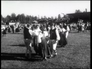 Dansande ungdomar i folkdräkt på gräsplan, läktare i bakgrunden.