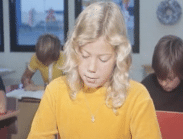 En sittande ung flicka med gul tröja som blickar ner på något i ett klassrum, ett par skolbarn i bakgrunden.