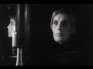 Kortklippt ung kvinna med höjd blick, hennes ansikte syns även i profil i en spegel till vänster i bild bakom ett brinnande stearinljus.