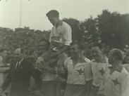 En fotbollsspelare med en pokal i famnen sitter på en kamrats axlar omgiven av fler leende medspelare, publik på läktare i bakgrunden.