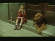 En liten flicka sitter i en port med en hund i koppel och spelar munspel, bredvid de båda står en liten miniatyrfigur.
