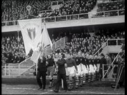 Två fotbollslag tågar in på plan efter ett par män med flaggor, publik på läktare i bakgrunden.