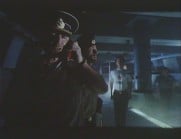 Mörk bild där en kapten på en u-båt svarar i telefon, i bakgrunden syns ett par besättningsmän.