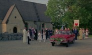 Ett bröllopspar sitter i en röd bil utanför en kyrka, ett tjugotal bröllopsgäster står runtomkring.