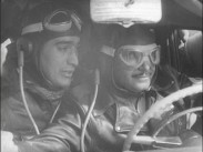 En rallyförare och en kartläsare med läderhuvor och skyddsglasögon av äldre modell inne i en bil.
