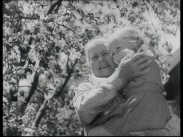 En äldre kvinna håller en liten flicka i famnen, blommande träd i bakgrunden.