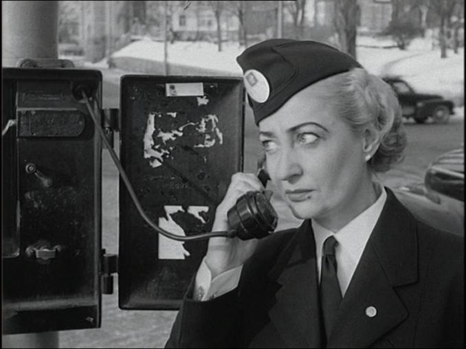 Uniformsklädd kvinna håller gammaldags telefonlur vid höger öra, bil i bakgrunden.