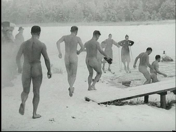 Fem nakna manliga vinterbadare på väg ner i en isvak vid en brygga, flera människor beskådar det hela.